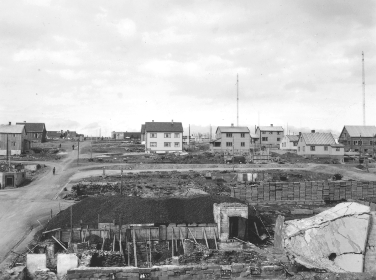 Motiv fra gjenreisningstiden i Vadsø etter andre verdenskrig. På bildet ser man flere nyoppsatte boliger. Nedenfor husene er det en stor ledig tomt. De to høye radiomastene til Vadsø Kringkaster ses mot himmelen