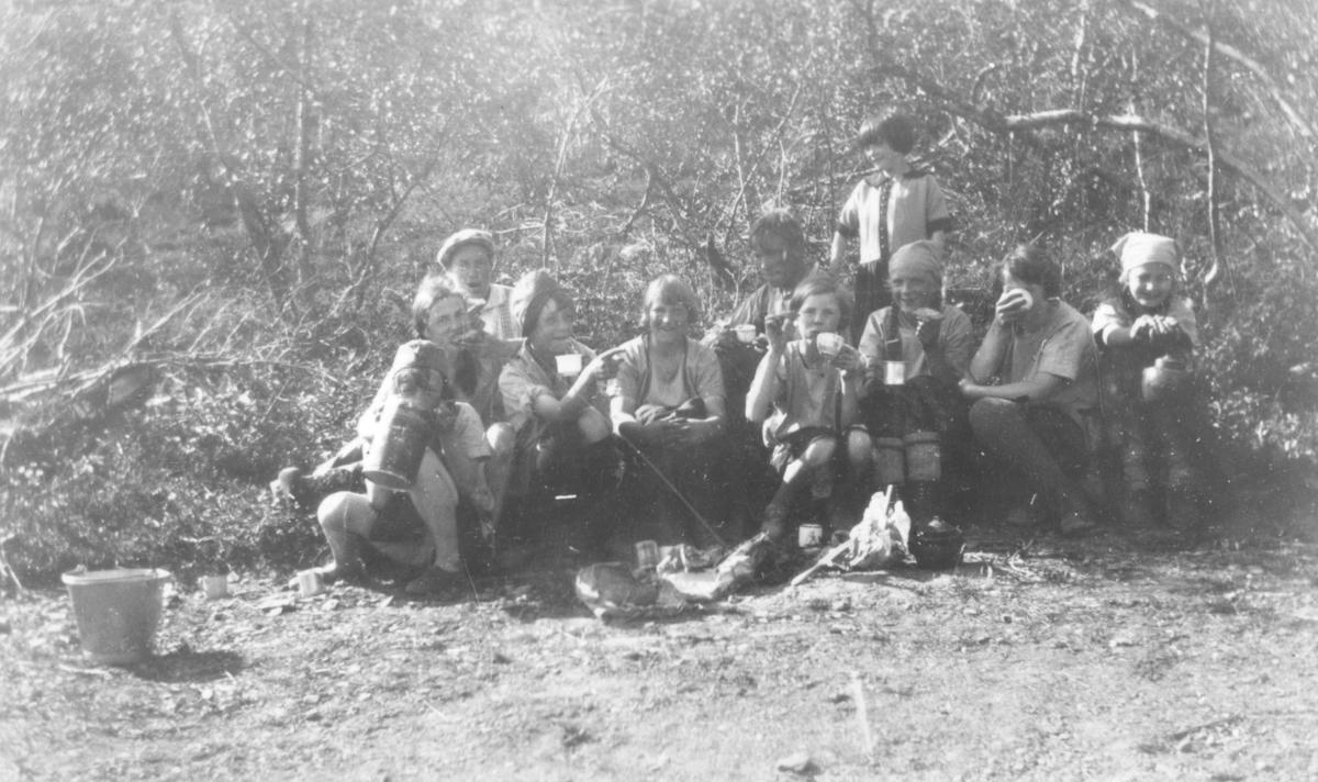 Kløverpatruljen på tur i Rustefjelbma, sommeren 1930. På bildet er de i torvmarka. I tekst til bildet står det ' Maten smaker godt i fjellet'. Vi ser de har matpause. Navn på noen av jentene på bildet er: Arnhild Tøgersen, Anna Andersen, Aud Johannessen, Vera Hoel, og Astrid Pedersen. Det er skog i bakgrunnen av der jentene sitter. Det står en bøtte på bakken ved siden av dem