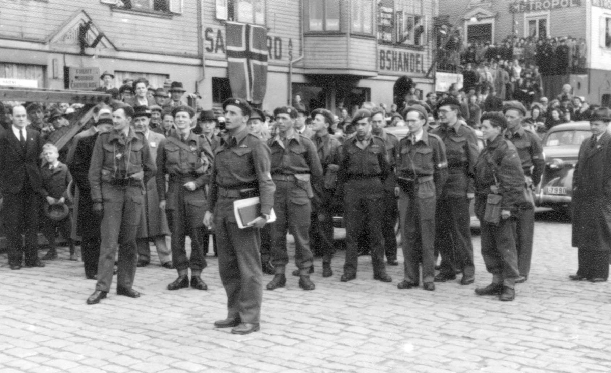 Frigjøringen i 1945 - opptog på indre kai. En gruppe soldater oppstilt foran et par biler. Gruppen er bare en del av noe som skjer lenger nord på kaien. Langs veggen og oppover i trapper og i vinduer er det fullt av folk som venter på at noe skal skje. Midt i bildet henger et norsk flagg ut av et vindu.