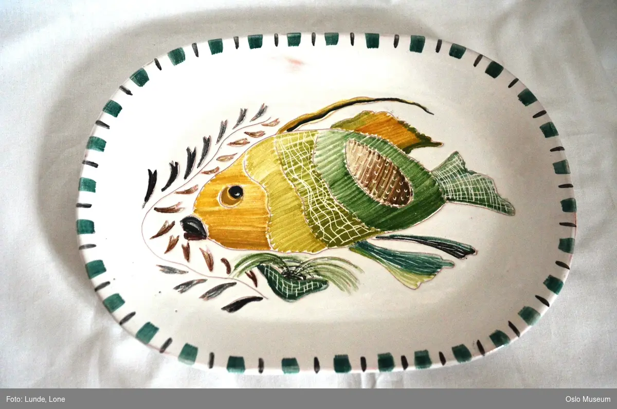 Avlangt fat i glasert keramikk. Hvit bunnfarge, grønn dekorkant, motiv i form av en fisk.