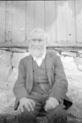 En eldre mann sittende foran en husvegg.