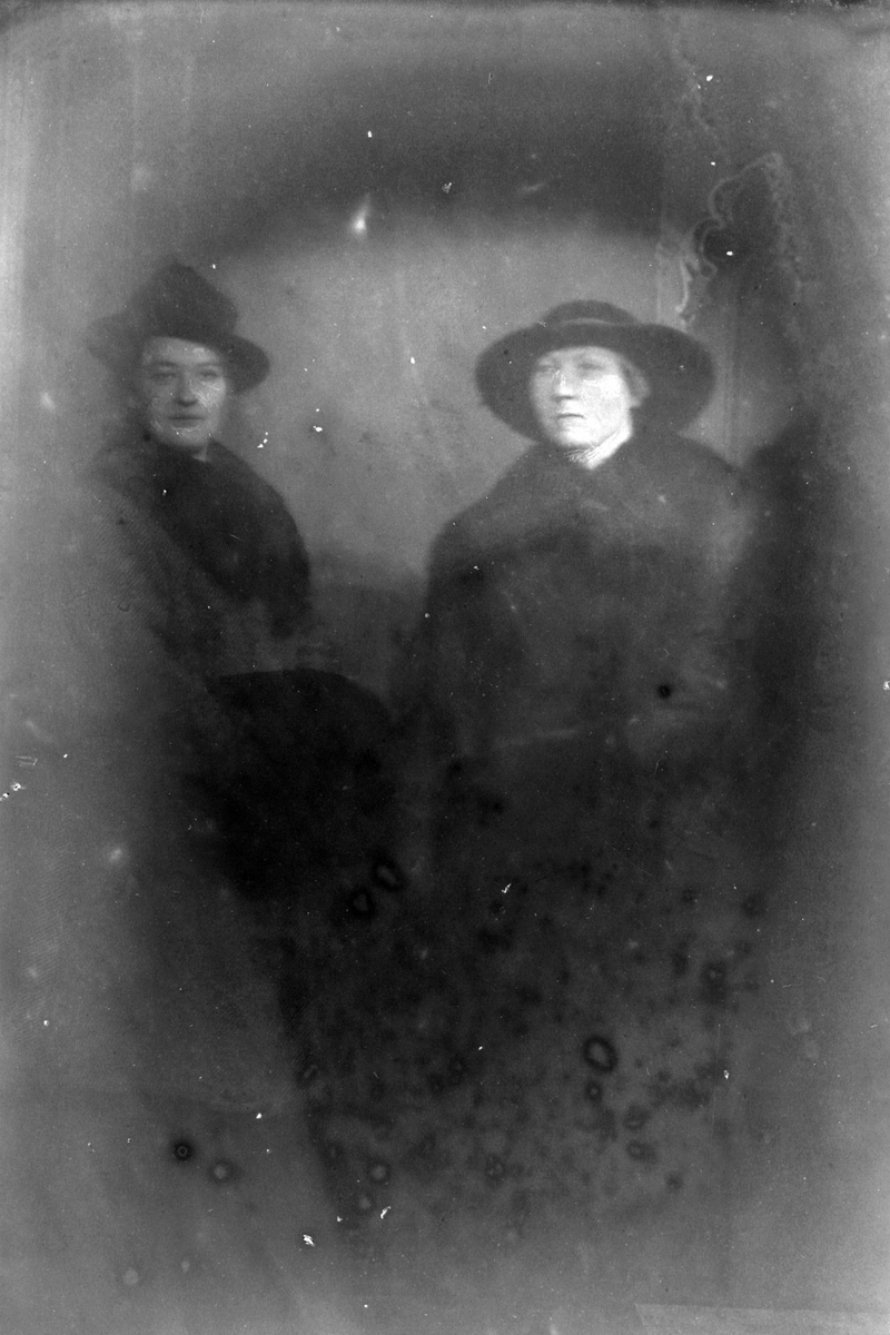 Studioportrett av to kvinner med hatter på hodet. Bildet er ganske ødelagt.