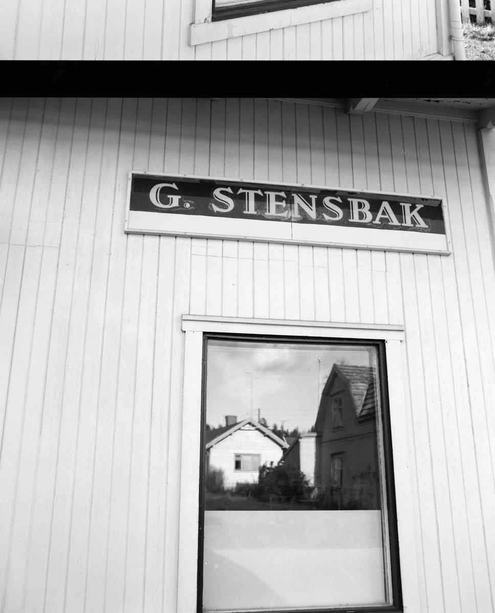 Butikkvindu, skilt med G. Stensbak(slakteforretning og pølsemaker. Klevbakken 83, Ådalsbruk. Bebyggelsen på andre sida speiler seg i vinduet. Løten.