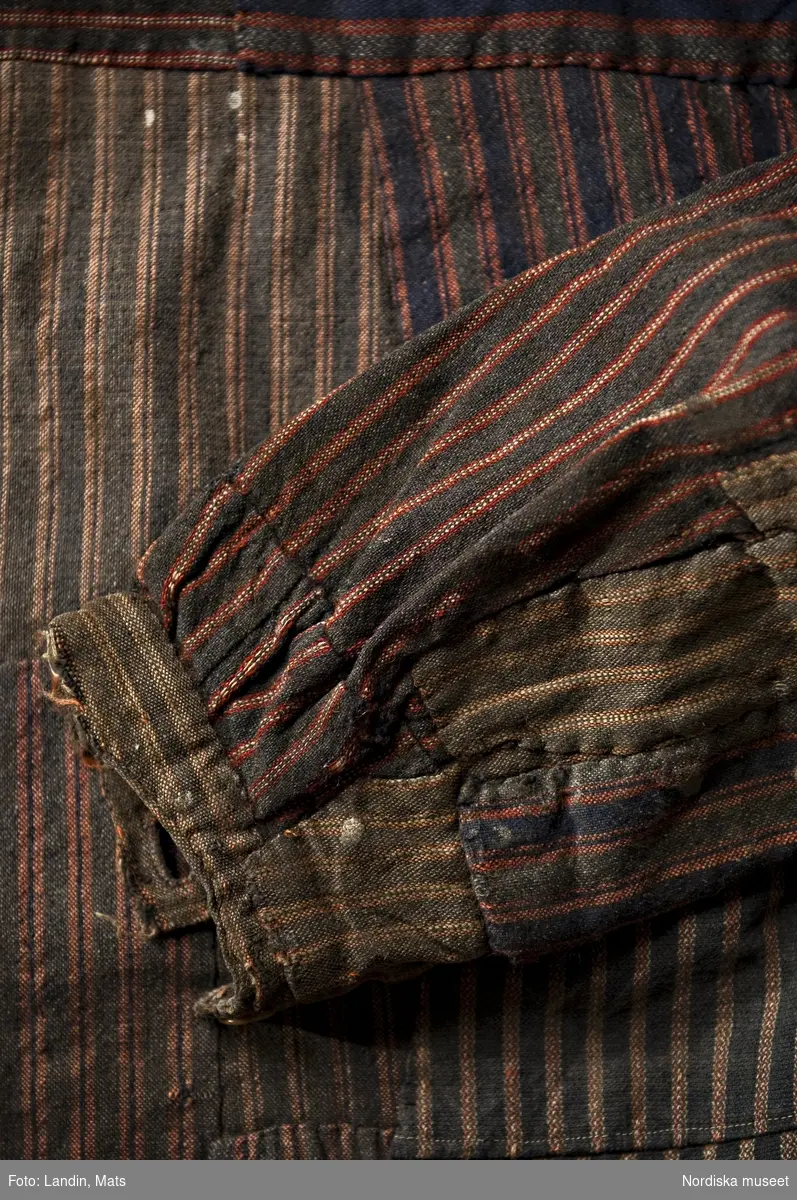 Skjorta, bussarong, arbetsklädsel, Uppland, Gräsö, 1900-talets början. Halvylle. 
Arbetsskjorta s.k. bussarong av hemvävt halvylletyg. Mörkblå bomullsvarp och inslag av ullgarn i mörkgrått och mörkblått med röda ränder och i vissa partier även en vit tråd. Traditionell skjortmodell med vidsydda ärmar med ärmspjäll (fyrkantig isättning) och ärmlinning. Skjortan är lappad upprepade gånger med totalt 19 lappar av olika randade tyger. Skjortan är maskinsydd, lapparna påsydda för hand, ofta på varandra. Den har använts ända fram till detta stadium av förslitning och blivit omsorgsfullt vårdad och lagad. Använd av fiskaren Karl Eriksson, på Gräsö, längst upp i Stockholms skärgård.