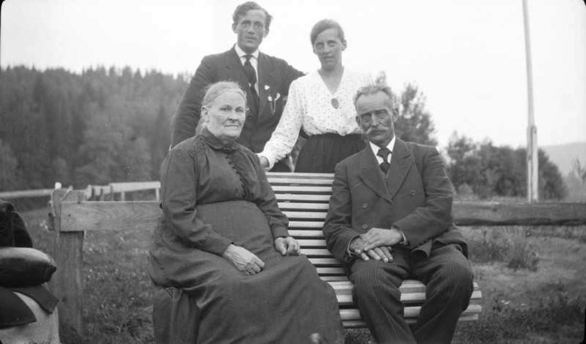 Et eldre par, Aagot Olausssens foreldre, på en benk.  Aagot Olausssen står bak  med sin bror.