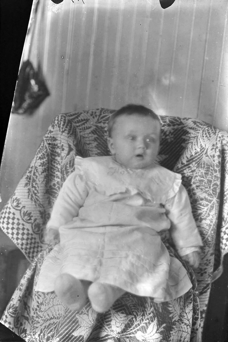 Portrett. Pikebarn/baby sitter på en stol dekket med et pledd som er mønstret. Barnet ser til side.Bildet er muligens tatt i Lofoten.
