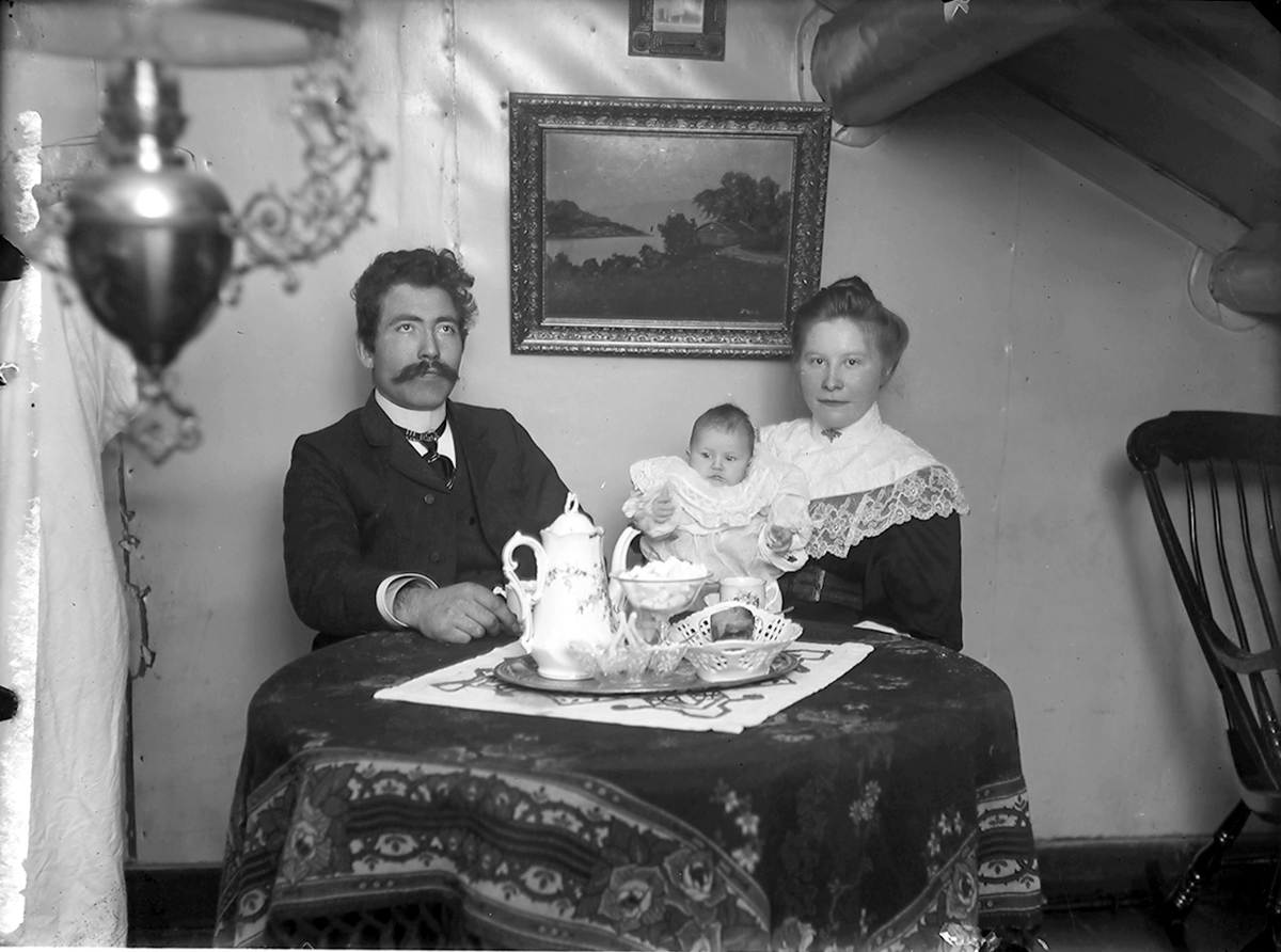 Portrett. Familiebilde. 3 personer i ei stue. Et par i 20-30-åra sitter ved et bord. På morens fang sitter et barn, ca. 1 år. Maleri på veggen. Lampe i venstre billedkant. På bordet et brett med kaffe og annet.Bildet er tatt i Glomfjord, Meløy. Interiør
