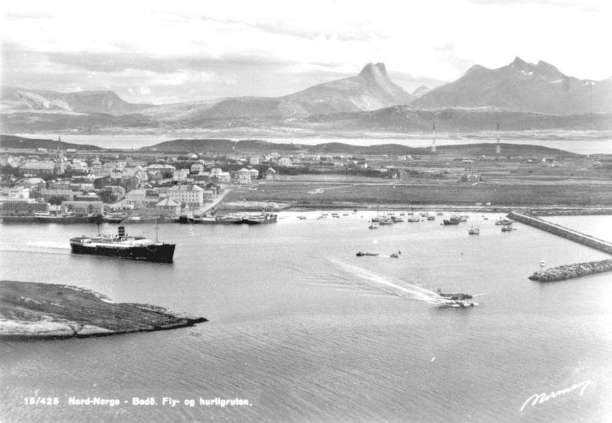 Utsikt over Bodø med havnen i forgrunnen. En hurtigrute og et sjøfly på vei ut av havnen. Moloen til høyre. Fjell i bakgrunnen.. 
Børvastindene i bakgrunn.