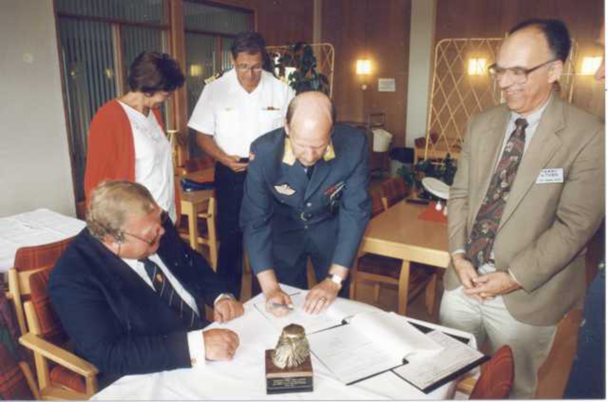 Fem personer ved et bord, en kvinne og 4 menn. Noen i militæruniform. Undertegning av kontrakt/avtale.
