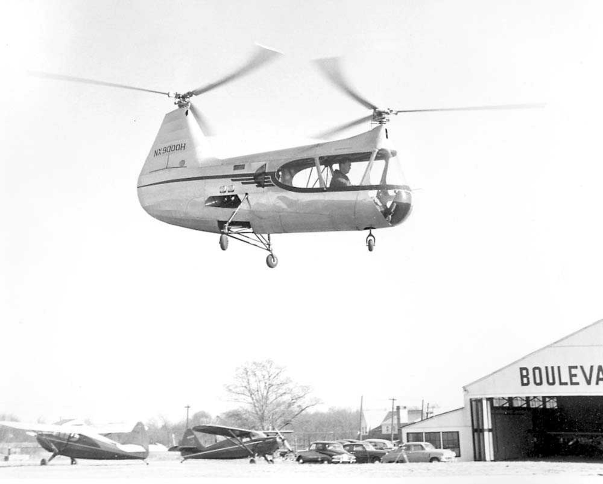 Ett helikopter i luften. HERC Jov-3. En person i copiten. Fly, biler og bygninger på bakken.