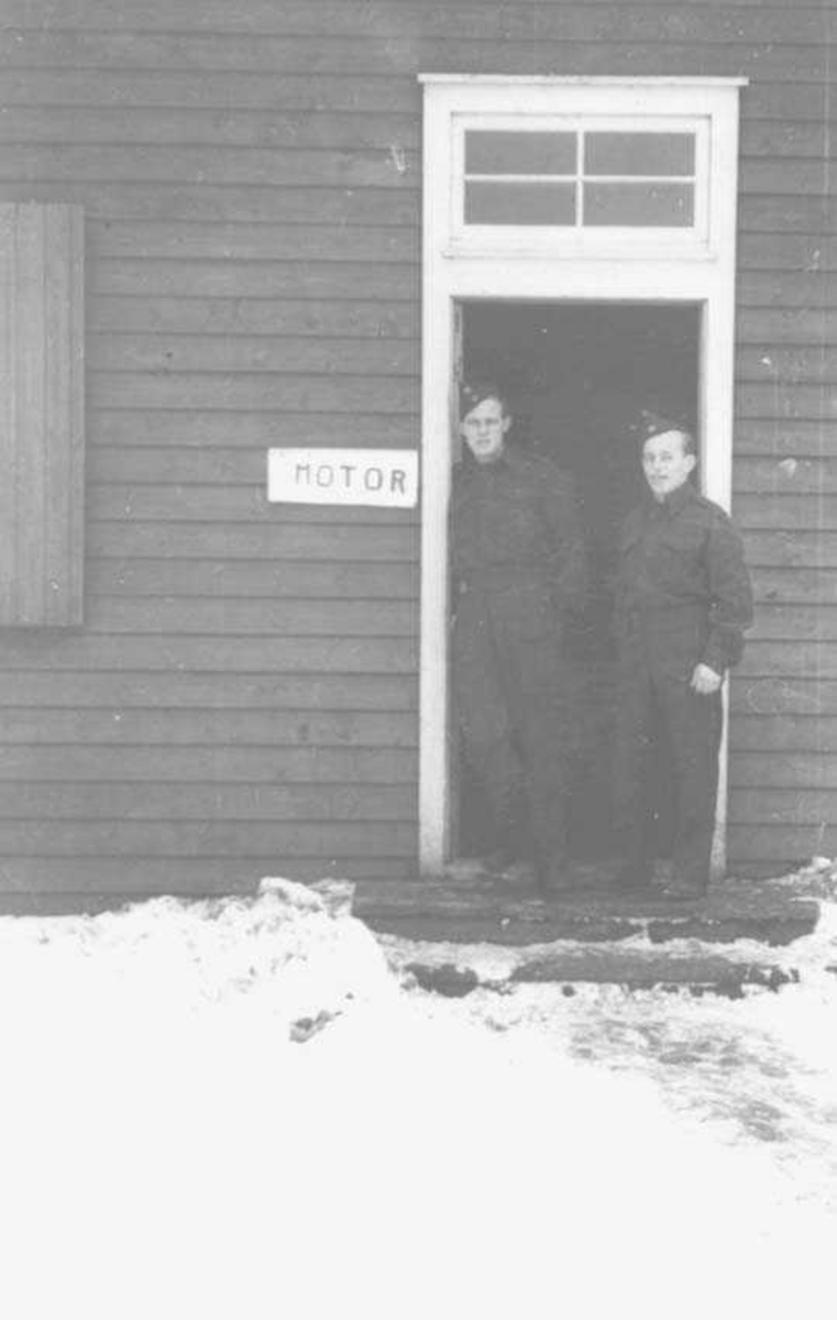 To personer som står i døråpningen til en bygning. Menn i militæruniform. Ved siden av døren er det et skilt som det står "motor" på.