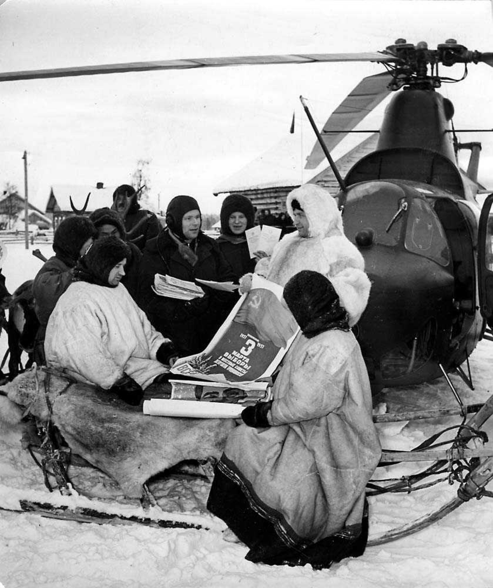 Gruppefoto. Åtte personer, to sitter på en slede, en deler ut politisk propaganda. Ett helikopter i bakgrunnen Mil Mi-1. Snø på bakken.