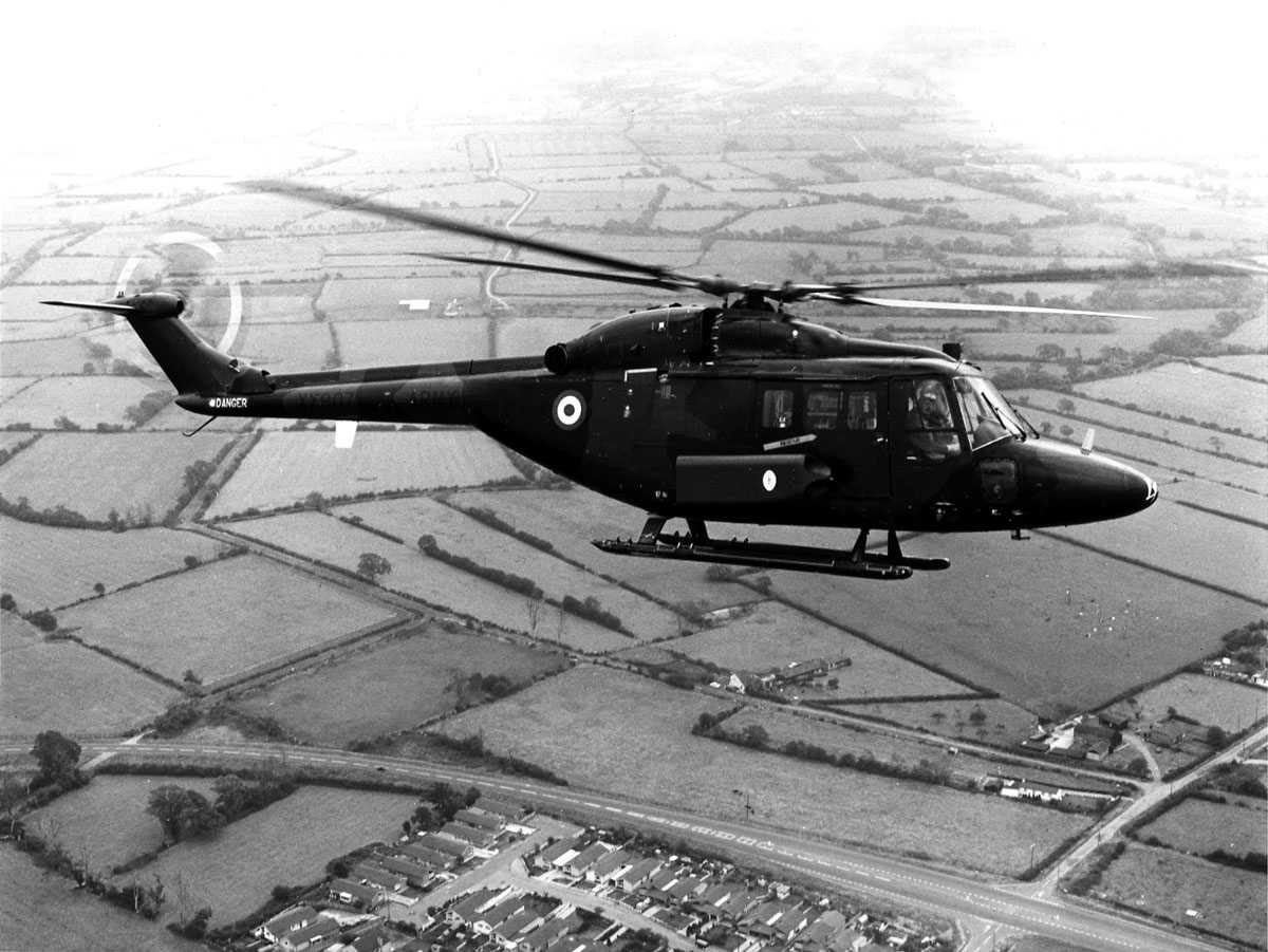 Etthelikopter i luften over jordbruksområder/bebyggelse, Westland Lynx AH Mk.1 tilhørende British Army merket XX907. 