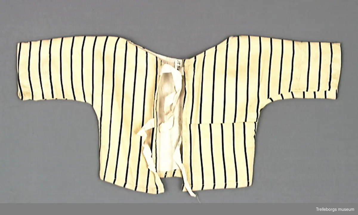 Babyskjorta med långa ärmar i ett gult tyg med svarta ränder. Skuren i ett stycke så det finns inga axel eller ärmsömmar. Halsringningen är rund och helt öppen framtill, den knyts ihop med två par bomullsband. Hela skjortan är fodrad med ett vitt bomullstyg.