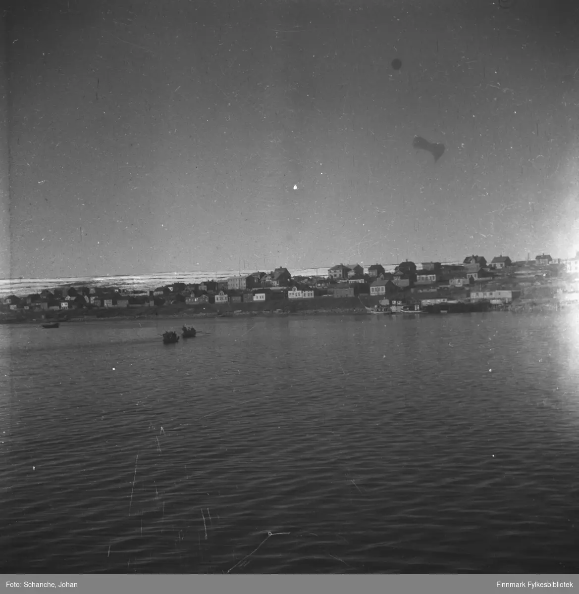 Indrebyen i Vadsø fotografert fra havet. To båter ror mot byen. Ruiner bland byggninger. Fjellen bak byen er delvis dekt av snø.