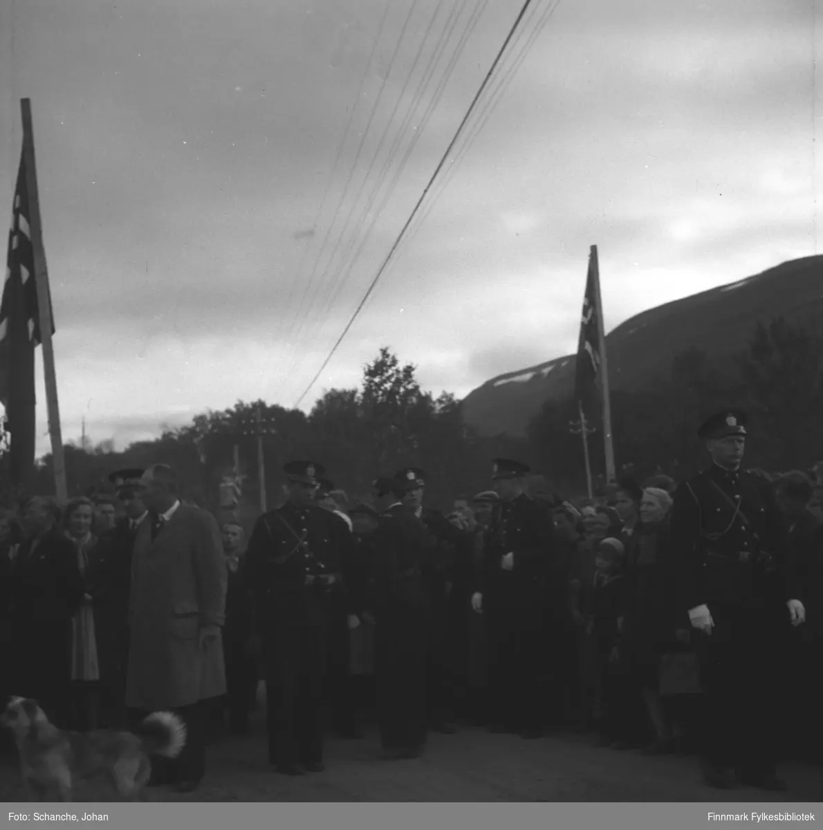 Kongebesøk i 1946:  Kong Haakon VII besøker Tana. Folkemengde står på gata foran Tana Turiststasjon og venter på Kongen. Fem politimenn i uniformer står fremst i mengden.