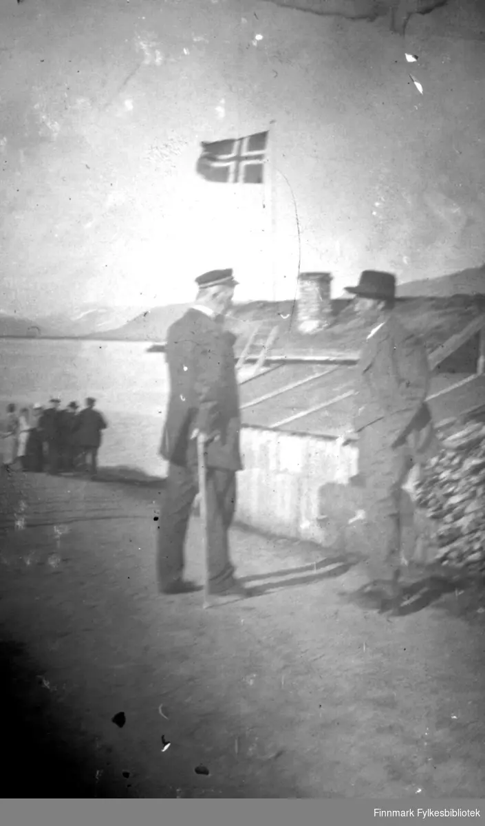 To menn i passiar på Langnes foran et lite hus med flagg. I bakgrunnen en gruppe mennesker. Mannen til venstre på bildet var lensmann i Tana fra år 1900 til 1935. Han hette Johan Kristian Mostrøm Bertheussen. Var født i Vardø i 1864