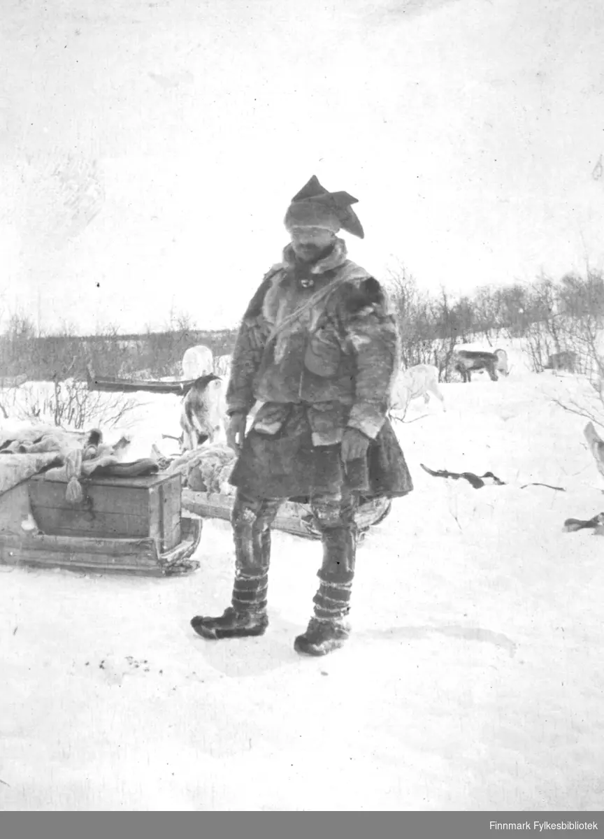 En mann, John Varsi, er fotografert ute i snøen. Han er kledt i pesk, stjernelue og skaller. I bakgrunnen er det reinsdyr og flere pulker.