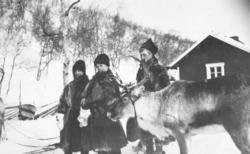 Tre personer og et reinsdyr fotografert utenfor et hus ved L
