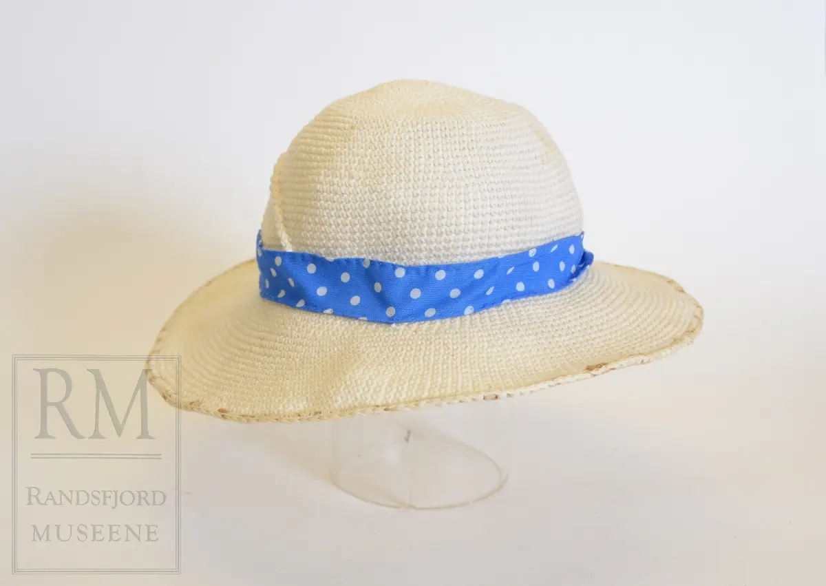 Sommerkjole m/hatt. a) Bomullstoff, blått med hvite prikker. Sammensydd i livet. Overdelen har Lukning foran med 5 hvite knapper og knapphull, puffermer. Krage med heppekant. 6 Bredder i utskrådd skjørt. Lukning til venstre for midt foran. 
b) Belte med knapp og knapphull. 
c) Hatt, Heklet av hvitt bomullsgarn. Ståltråd ytterst i Bremmen (rusten) Figurkant på pullen. Pyntebånd av kjoletøyet.