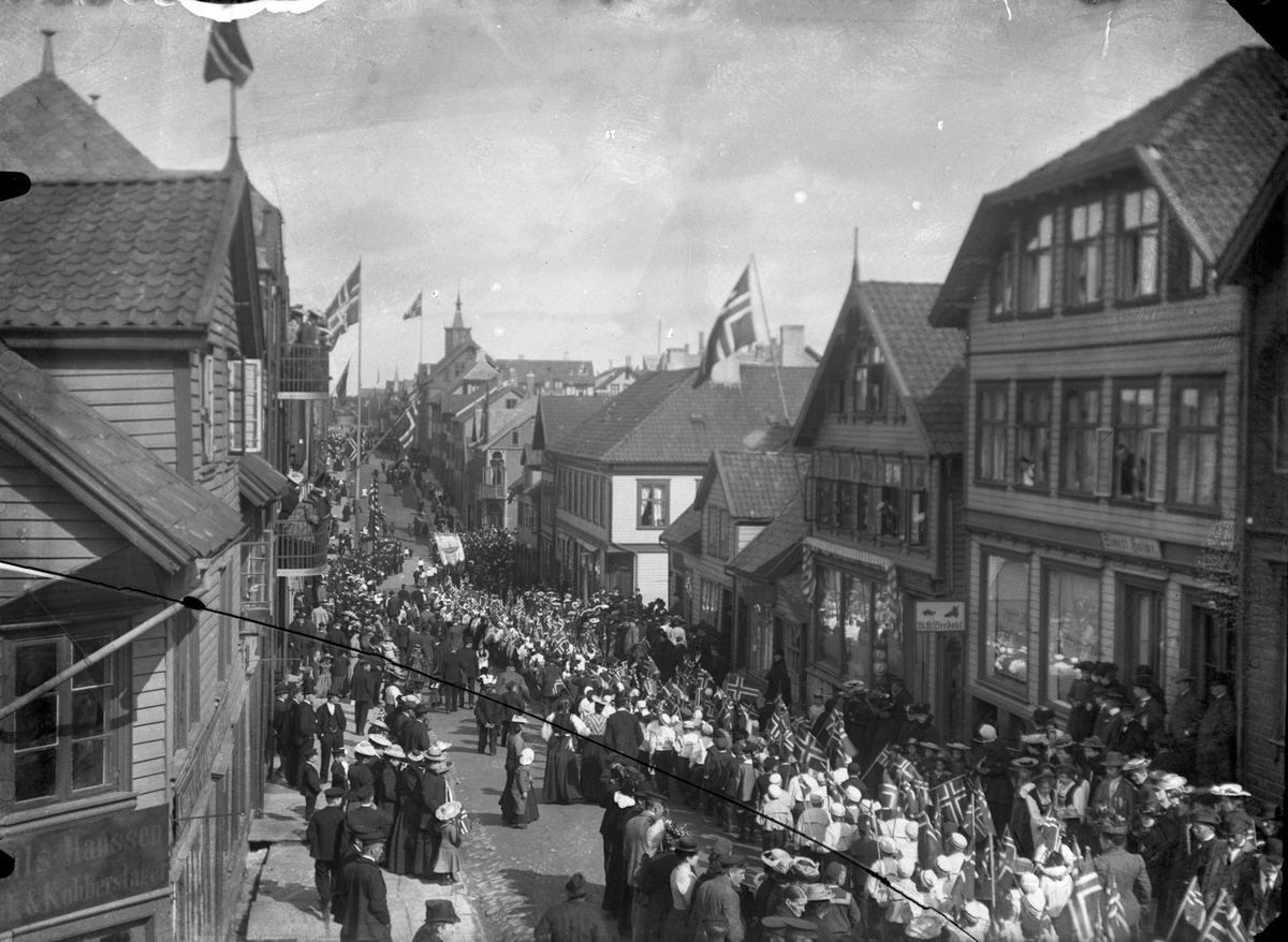 17. mai tog i Strandgaten. Gaten full av barn med norske flagg. De fleste voksne står på fortauet. Trehus på begge sider av gaten. I bakgrunnen er tårn og spir fra Vår Frelsers kirke synlig (?).