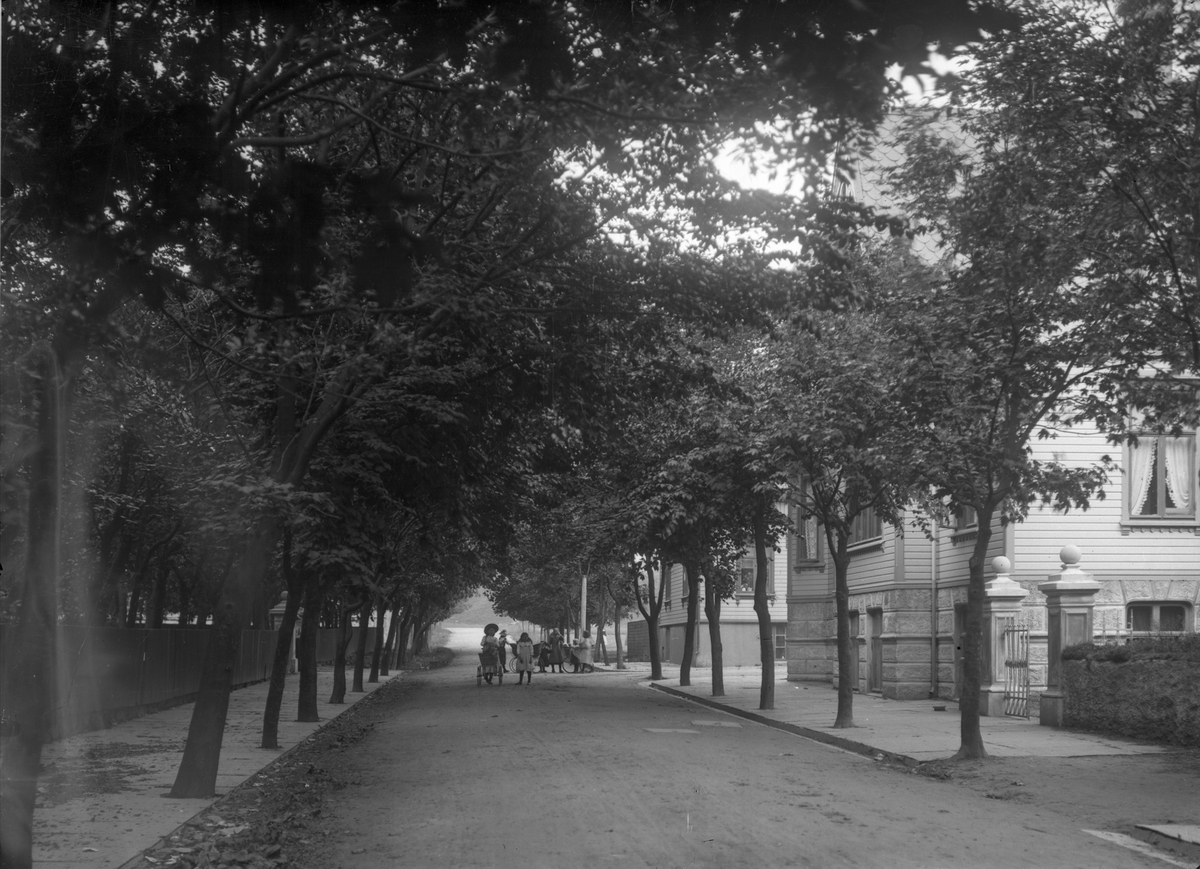 Byparken. Trær til venstre, hovedsaklig trehus til høyre. Barn midt på bildet i gaten, delvis i bakgrunnen.  Fortau på begge sider.
