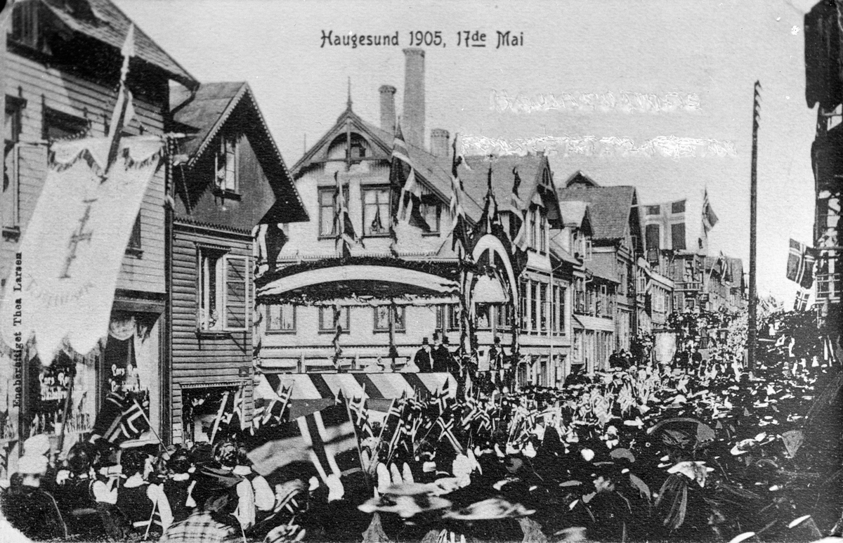 17 mai i Haugesund i 1905. Mange folk går i 17. mai tog. Flere norske flagg. En fane lengst til venstre, og i bakgrunnen. Flere personer til høyre på bildet.
