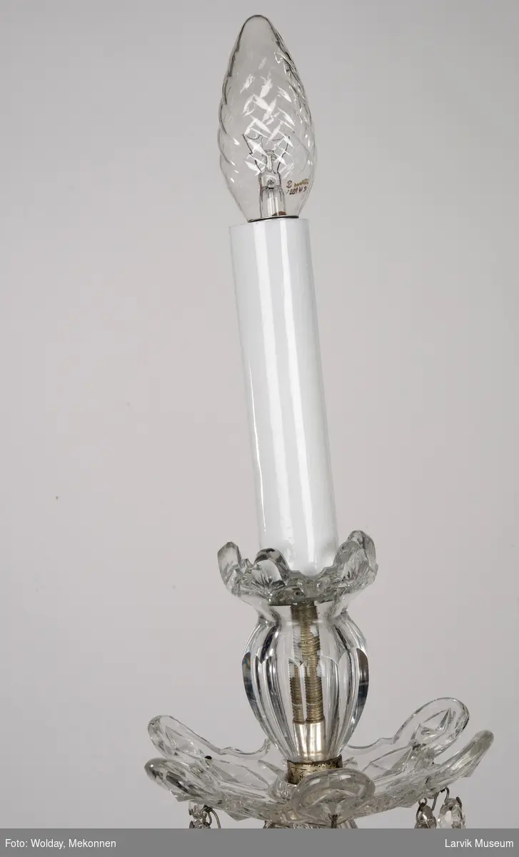 Balusterformet lysekrone.Slepen stamme med 6 svugne lyseholdere. Glassskål skjuler armfeste av stål, prismer i kjeder og enkle. Imitasjon av stearinlys i glass.
