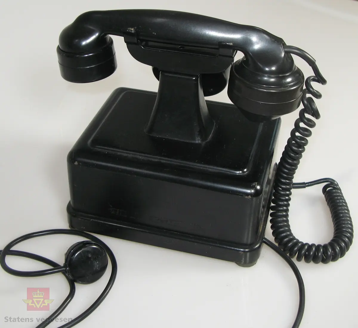 Svart telefonapparat. På fremsiden står det "Telegrafverket" sammen med logo. Apparatet har svart telefonledning med tre-polet kontakt.