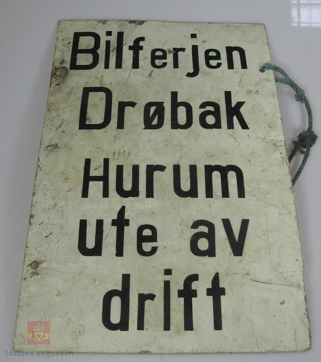 Rektangulært skilt av stål med svart tekst på hvit bunn. Tekst: Bilferjen Drøbak Hurum ute av drift. Teksten er teipet på skiltet. Skiltet har tre hull til bruk ved fastsetting.