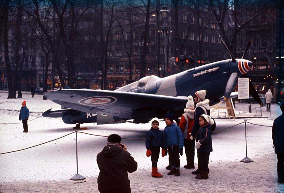 Ett fly som står utstillt i en park, Spitfire Mk P. R. XI AZ-B. Flere personer rundt flyet, bygninger i bakgrunnen. Snø på bakken.