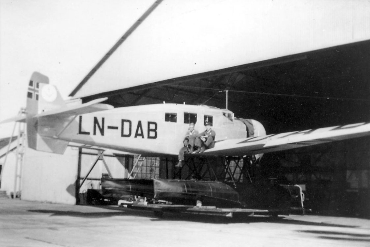 Ant. sjøflyhavn. ett fly på bakken, Junkers Ju W 34 "Ternen", LN-DAB, foran en hangarbygning. To personer sitter på den ene vingen.