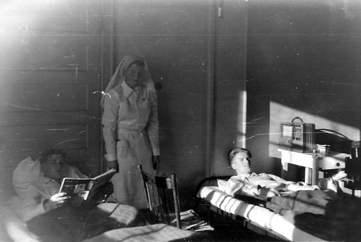 Portrett, tre personer hvor to er mannspersoner som ligger i senger, ant. sykesenger. I midten står en sykesøster..