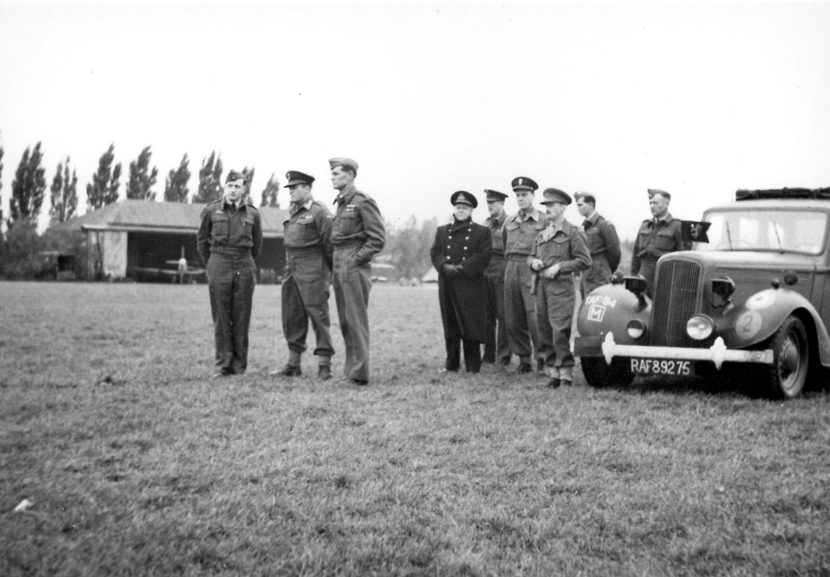 En gruppe personer i militæruniform, tatt utendørs. Kronprins Olav og en gruppe personer. T.h. bil med regisrering RAF 89275. Bak sees et fly inne i en hangar.