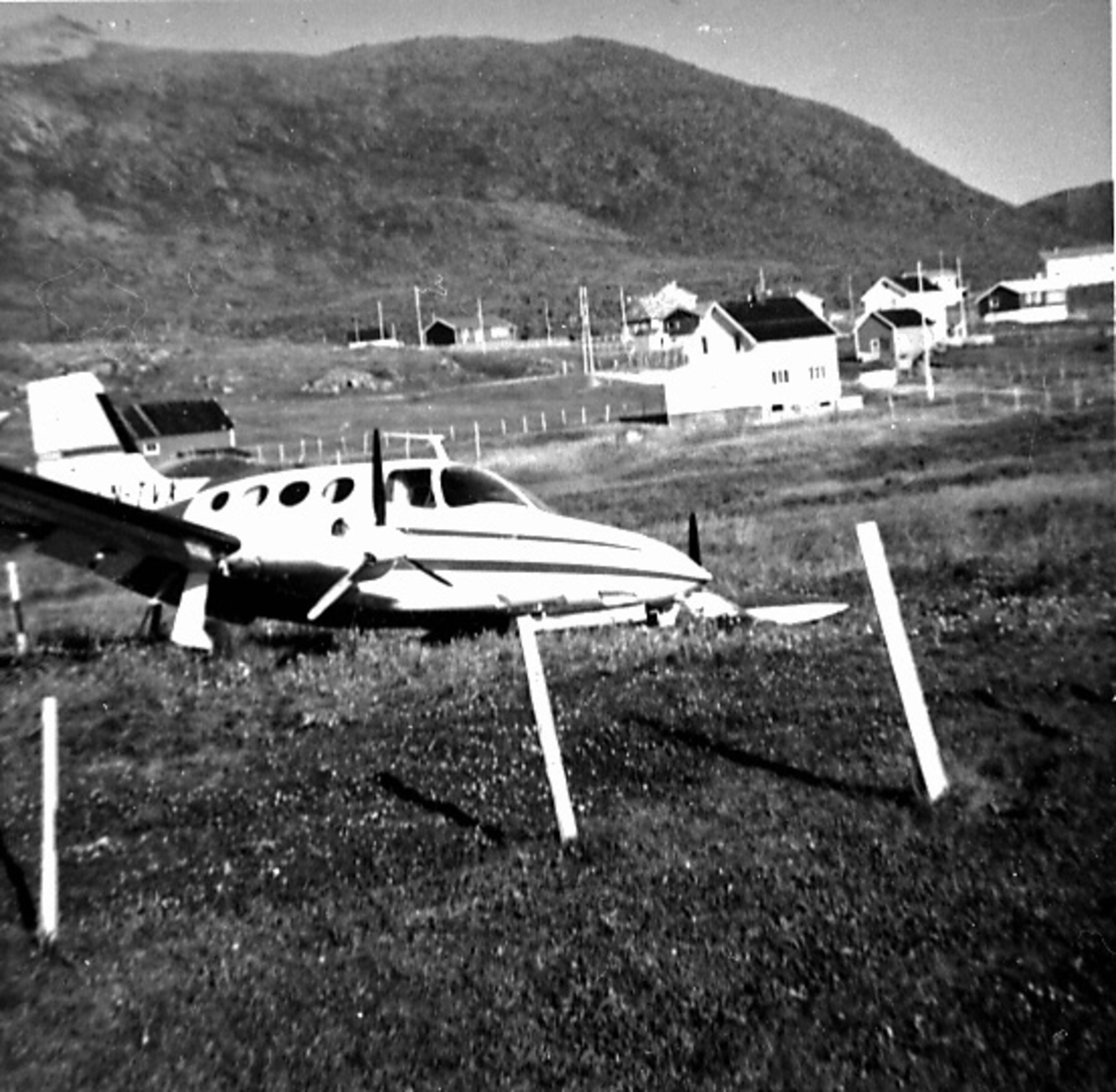 Fly, Cessna 401A, LN-TVX. Skrått forfra. "Ligger" ute på et jorde. Ene vingen nede i bakken. Bygninger bak.