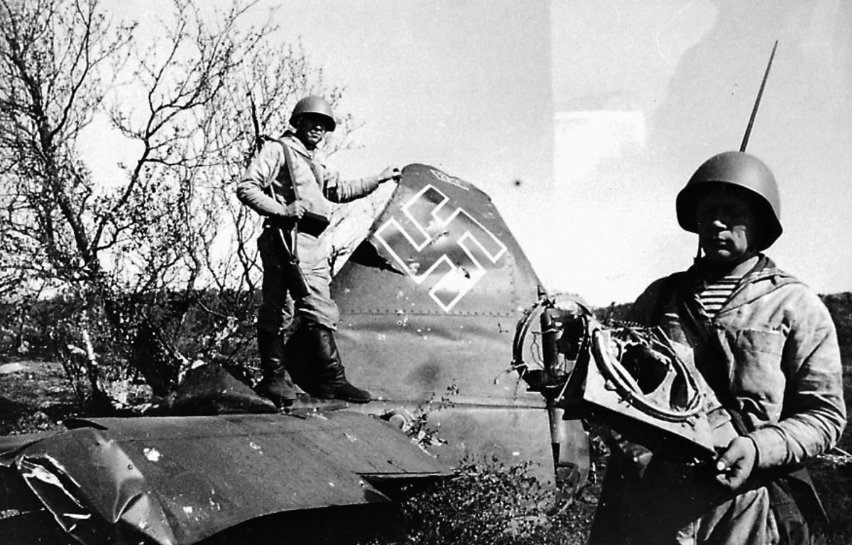 Portrett. 2 soldater i militæruniform. Begge med gevær på ryggen. Står ved et ødelagt fly, Ju 88 A-5 (W.Nr.2341, 4D+AC?). Hakekors påmalt halepartiet.