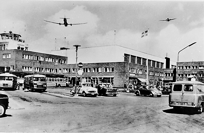 Lufthavn. Fra innkjørselen, Flere kjøretøy parkert, terminalbygningen bak og kontrolltårnet. 2 fly i lufta over bygningen.
