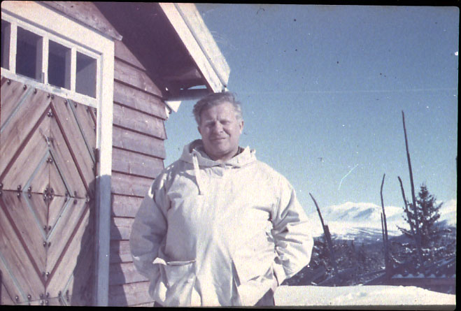 Portrett, 1 person i hvit anorakk, brystbilde, tatt utewndørs. Bygning t.v. Snø på bakken