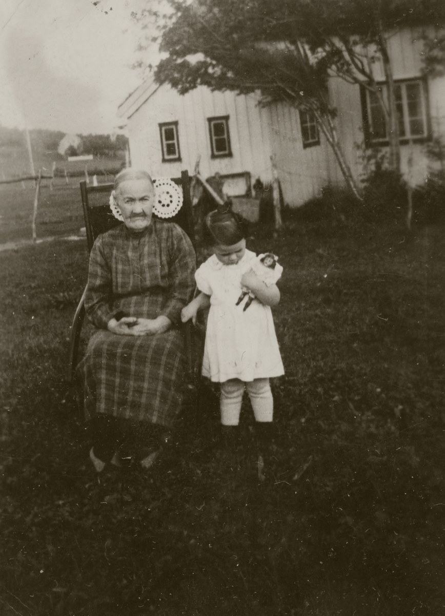 Eldre kvinne og lita jente med dukke, fotografert utendørs med et hus i bakgrunnen.