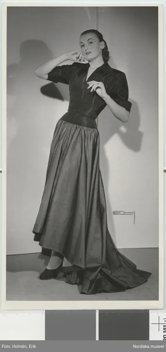 Nordiska Kompaniet. Kvinna i aftonklänning med svart liv och mörk nederdel som bakåt bildar ett litet släp. Text på baksidan: "Jean Desses uppvisning mars 1947."