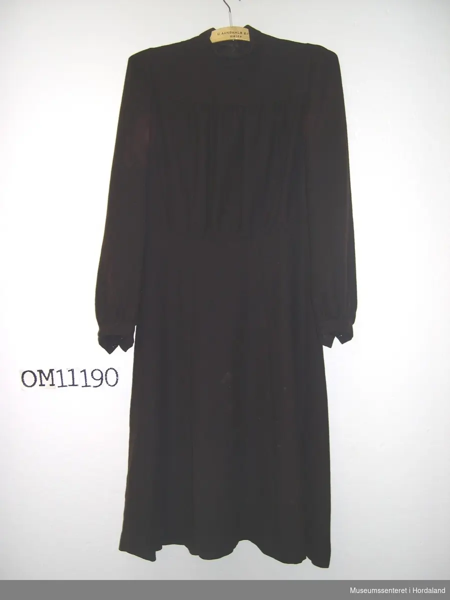 Form: langerma kjole med mansjettar, skrådd skjørt, sidelukking med trykk-knappar, og halslinning lukka med fire patentknappar bak  
