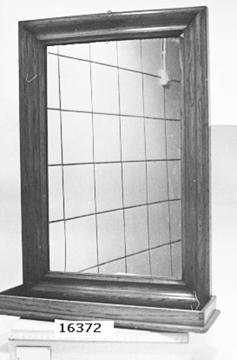 Rektangulär spegel med enkelt profilerad ekram. I underkant en hylla att lägga toalettsaker, rakdon med mera. Märkt på baksidan: kattfot, af Chapman.