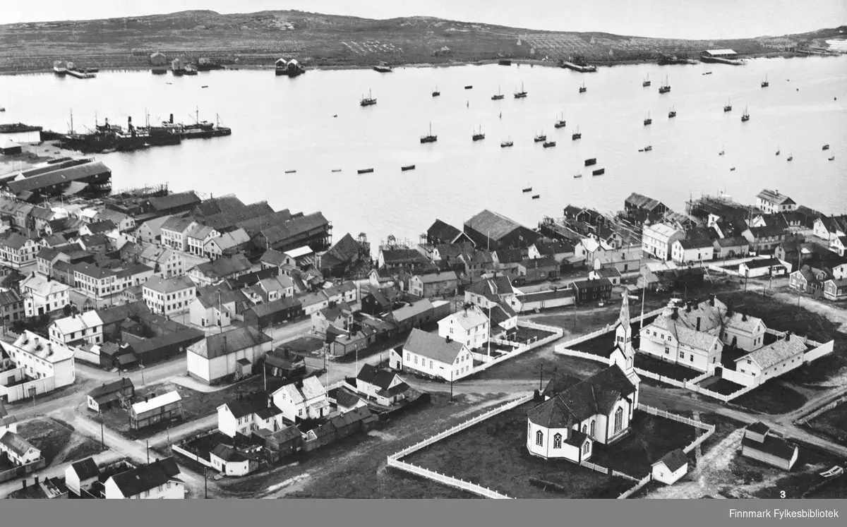Et postkort fra Vadsø. Foran på bildet kirke og kirkegården. Bakerst fjorden og øyen. Bildet er tatt fra radiomastene.