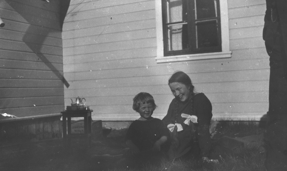 To jenter, "Biddi" Birgit Hermansen og Lillemor sitter utenfor dr. Kohls hus i Tana sommeren 1925. Sola skinner, det står en kaffekjele på et lite bord.