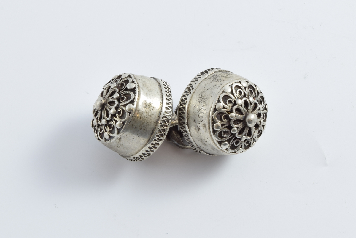 Halsknapper i sølv prydet med 8-bladet lokkerosett i en krans av 10 lokker. Mellom lokkene og i rosettens sentrum er det små perler.