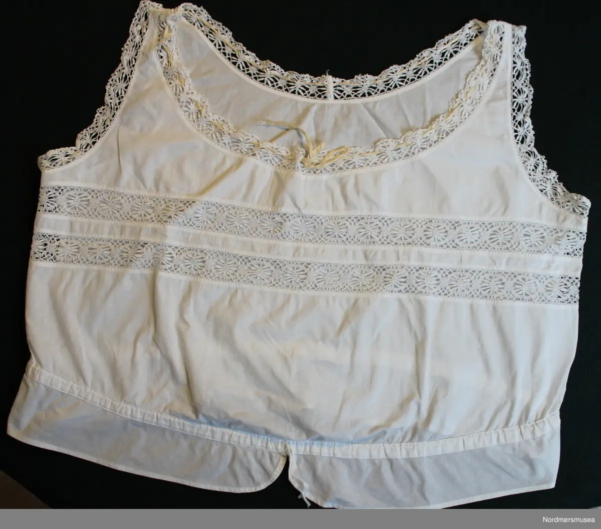 Liv/underskjorte for dame. Tynt bomullsstoff. Snøring i livet og ved hals. Innfelte blondebånd over brystet og ved avslutning ved armer og hals. Maskinsøm.