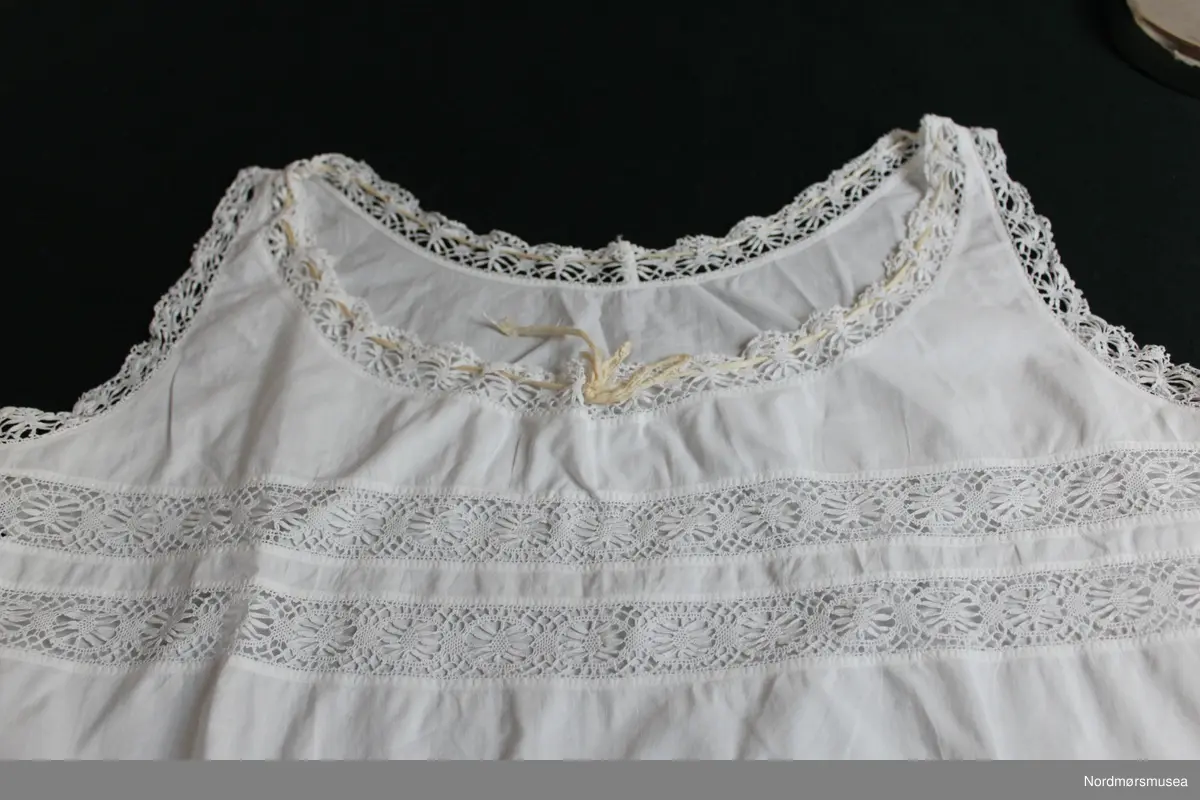 Liv/underskjorte for dame. Tynt bomullsstoff. Snøring i livet og ved hals. Innfelte blondebånd over brystet og ved avslutning ved armer og hals. Maskinsøm.