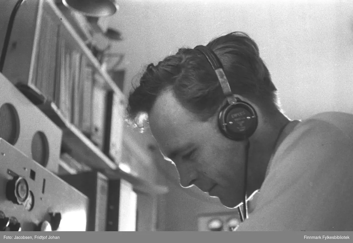 Aksel Jensen på jobb hos Telegrafverket (som det het til 1969) i Hammerfest. Han er iført en hvit skjorte og har hodetelefoner på seg. Foran han står noen apparater og en bokhylle med bøker/permer i.