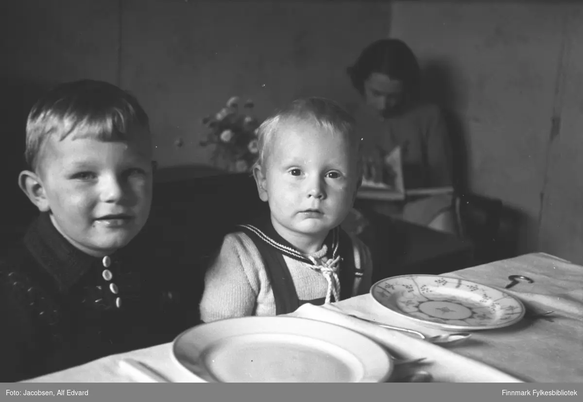 To små gutter sitter ved et bord. Det er fetterene Åge Christian Jacobsen til venstre og Arne Jacobsen. I bakgrunnen sitter Aase Jacobsen, mor til Arne. Åge har en mørk jakke med krage og lys knapper i halsen. Arne har en lys/hvit genser med mørk krage og snorer i halsen. Selene på en snekkerbukse går over skuldrene hans. Det er en hvit/lys duk på bordet og oppå står to tallerkener, den ene hvit og den andre med stråmønster. Noe bestikk ligger også ved siden av. Stolene de sitter i har mørke rygger og armlener. Aase har en ganske lys kjole på seg og sitter og blar i en bok. På bordet ved siden av henne står en blomsterbukett.