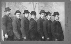 Gruppeportrett av syv menn tatt i et fotoatelier. Mannen som